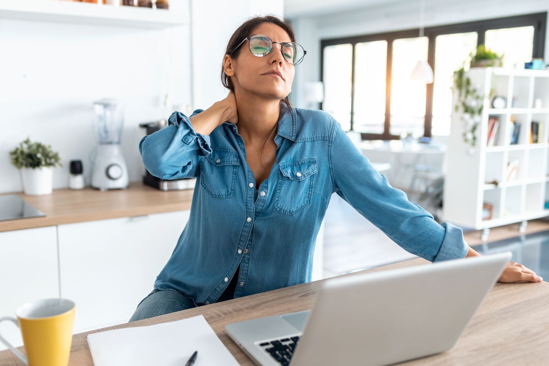A person with glasses wearing a blue shirt sits at a desk, rubbing their neck while looking exhausted.
