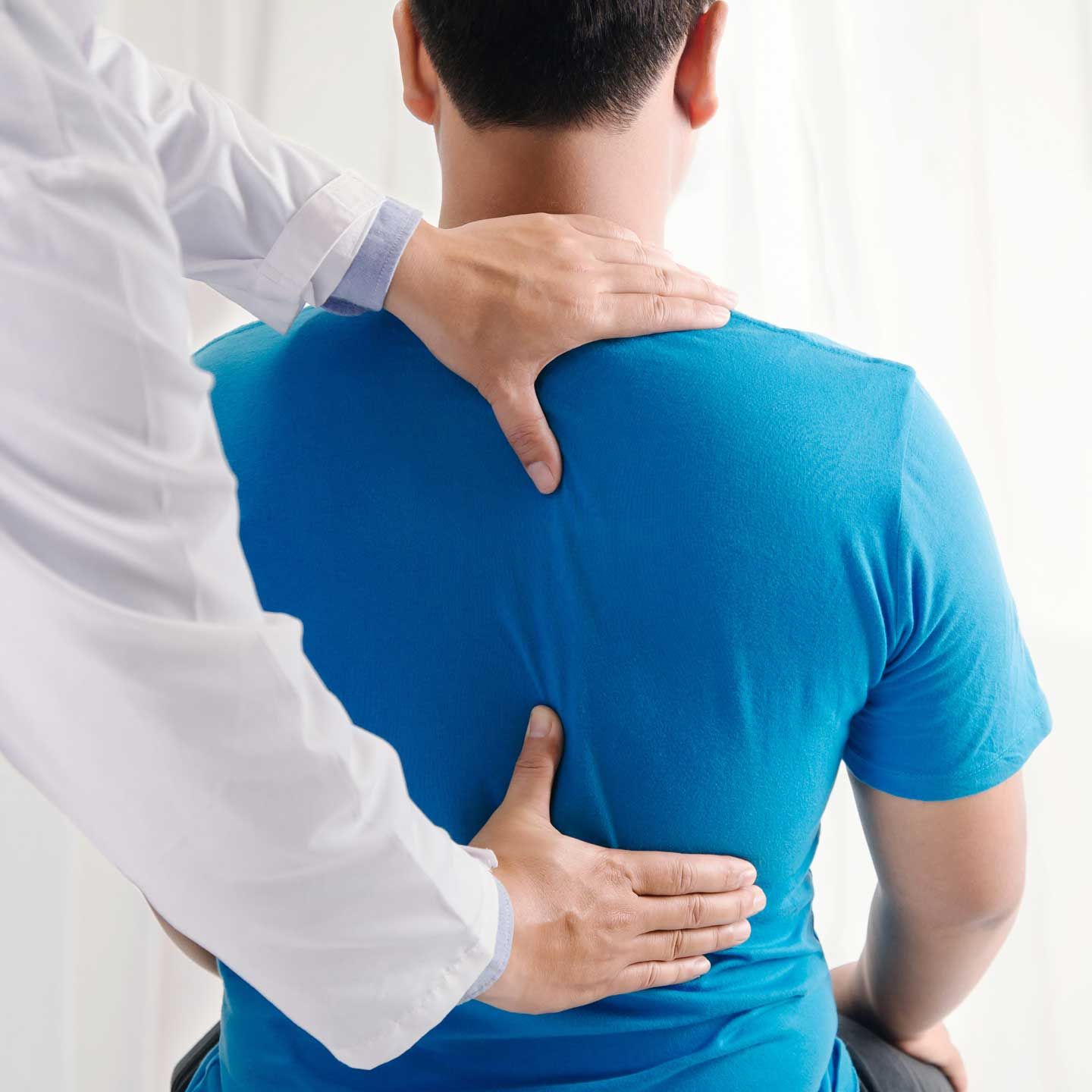 Doctor examining a patient's back. Hands on the patient's upper back. Blue shirt, white coat. Indoor setting. A professional in a white coat performing a back examination or physical therapy on a person wearing a blue t-shirt.