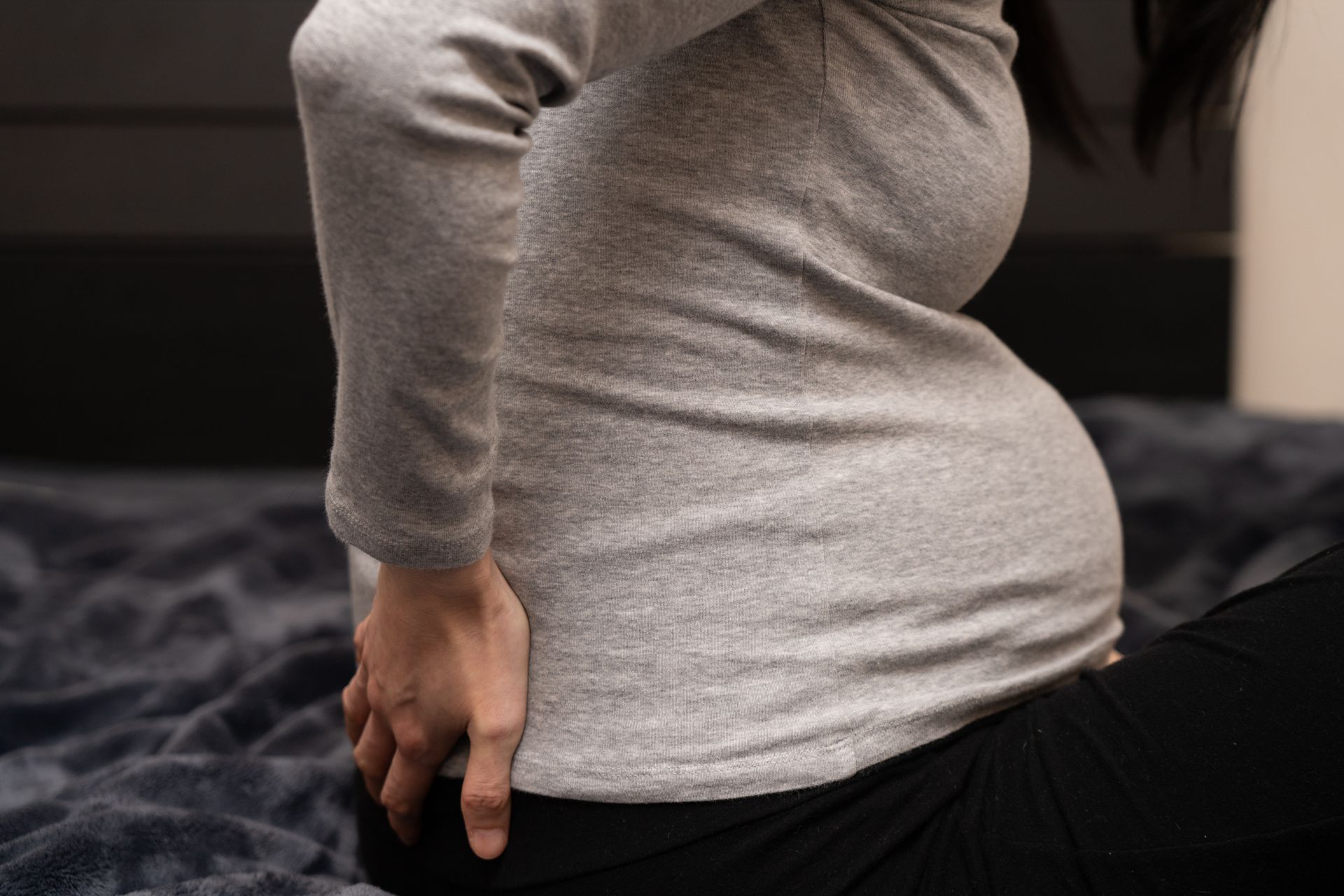 A person sitting on a dark bed, clutching their lower back with their hand, appearing to experience discomfort.