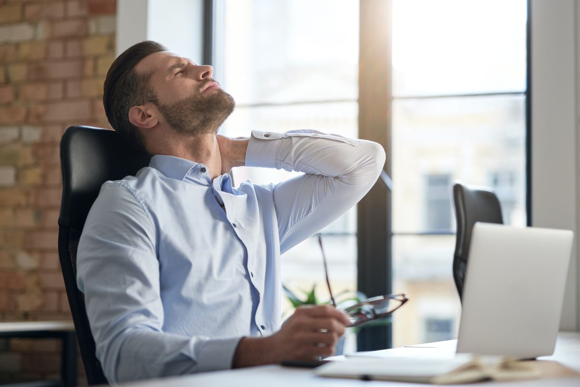 A professional sitting at a desk with a laptop, looking exhausted while holding the back of their neck.