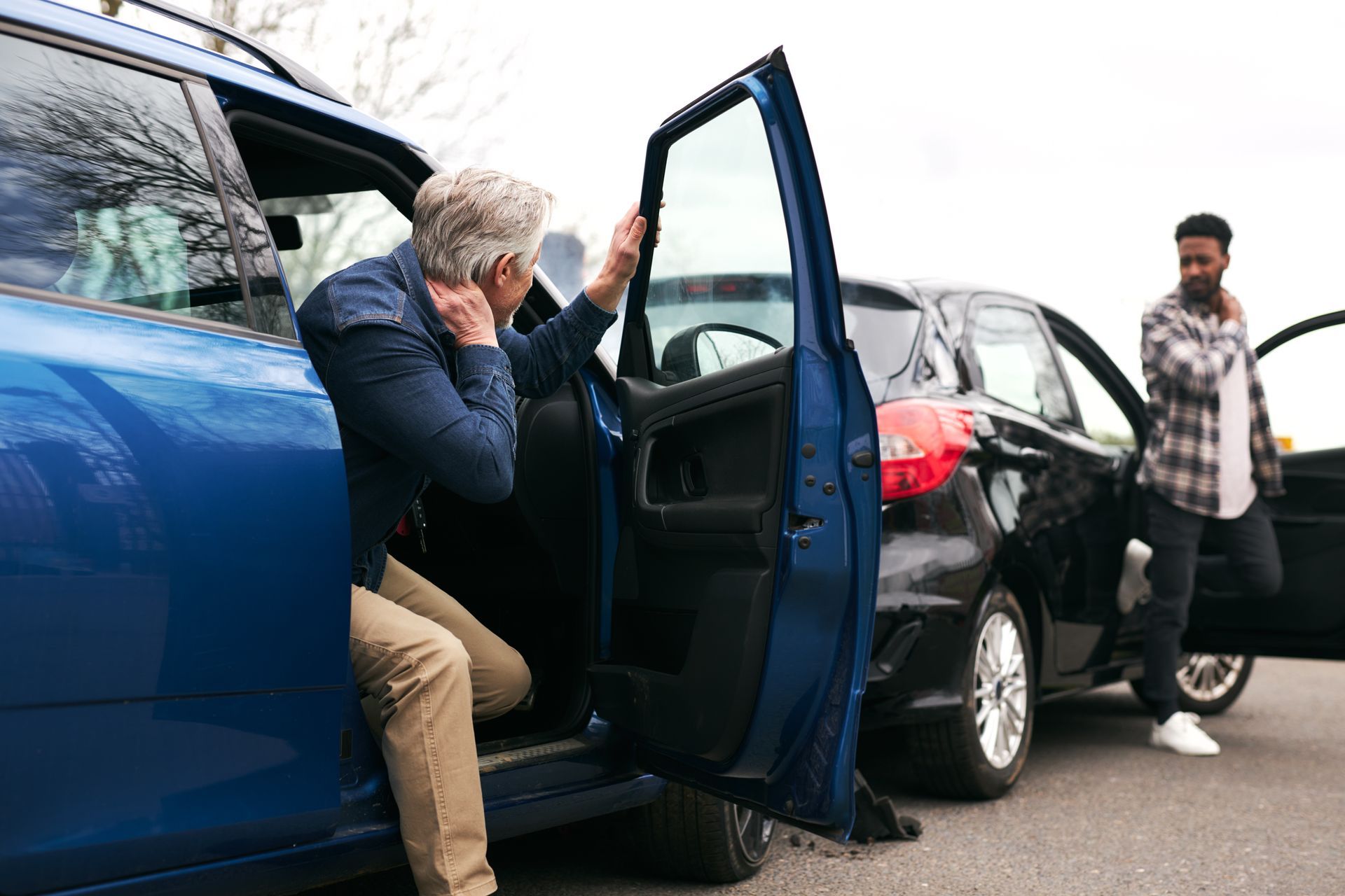 Two drivers step out of their damaged cars after a minor road accident, both holding their necks in pain.