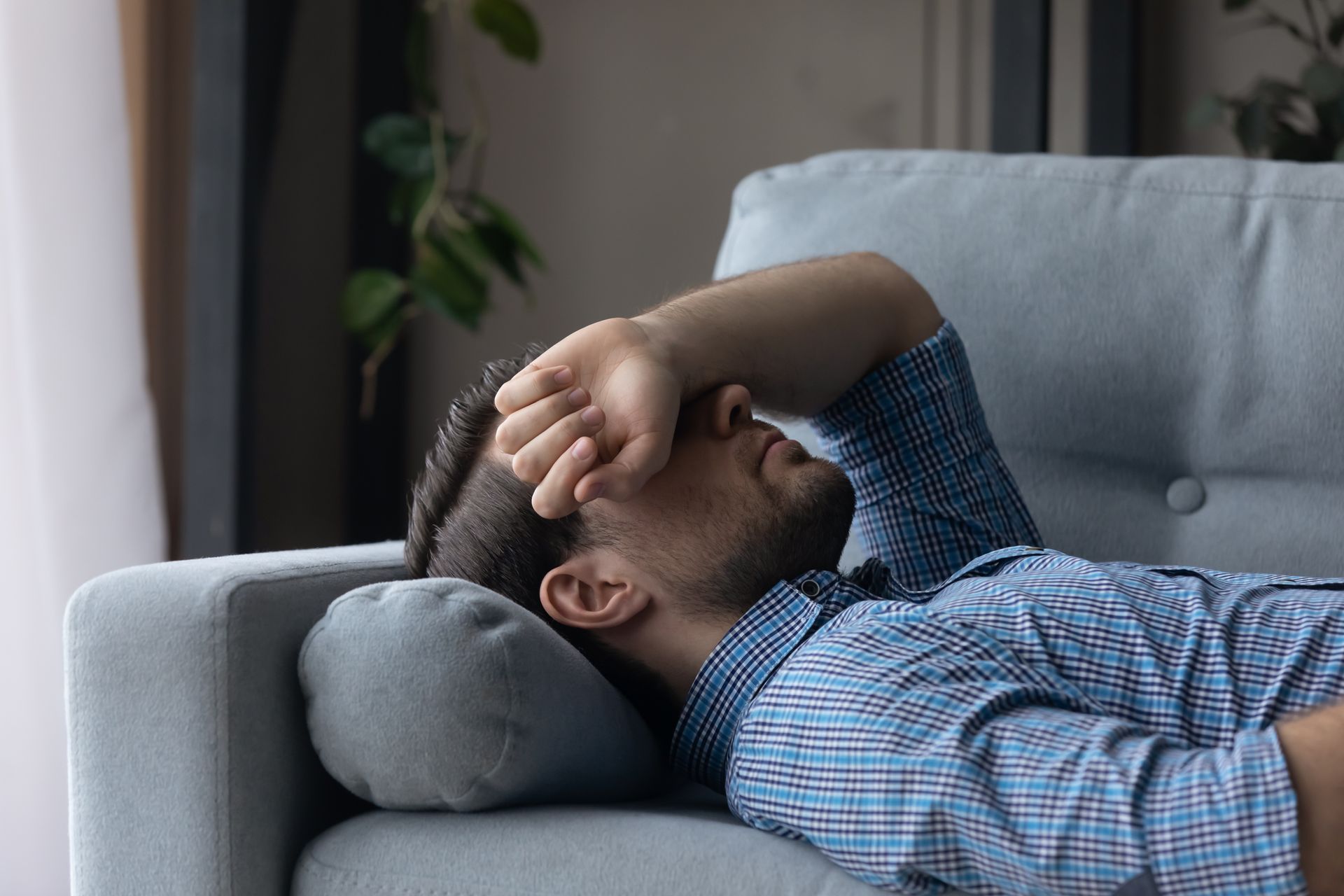 A person in a blue plaid shirt lies on a gray sofa with their hand over their face, appearing exhausted or distressed.