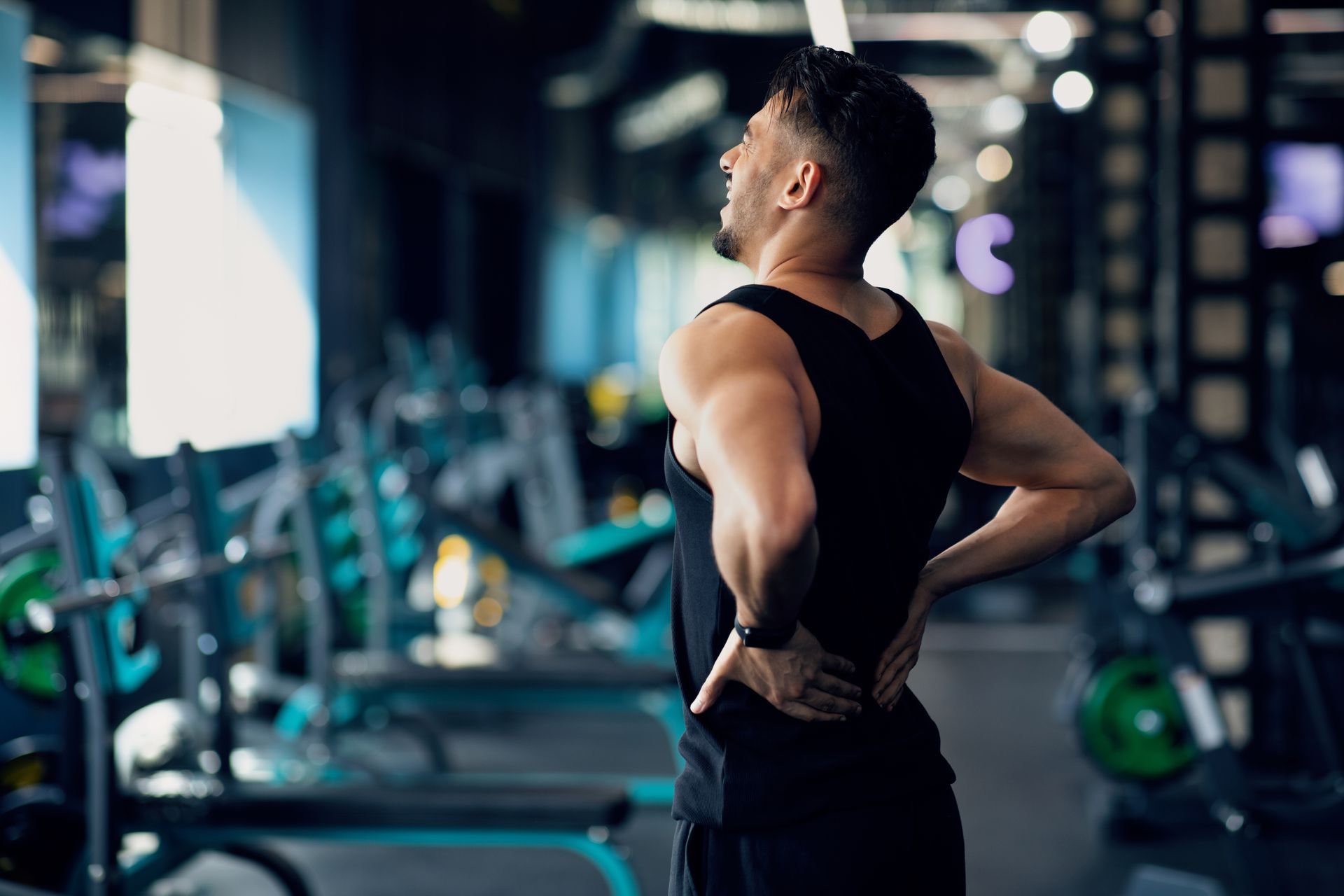A person in a black tank top stands in a gym, grimacing and holding their lower back due to pain.