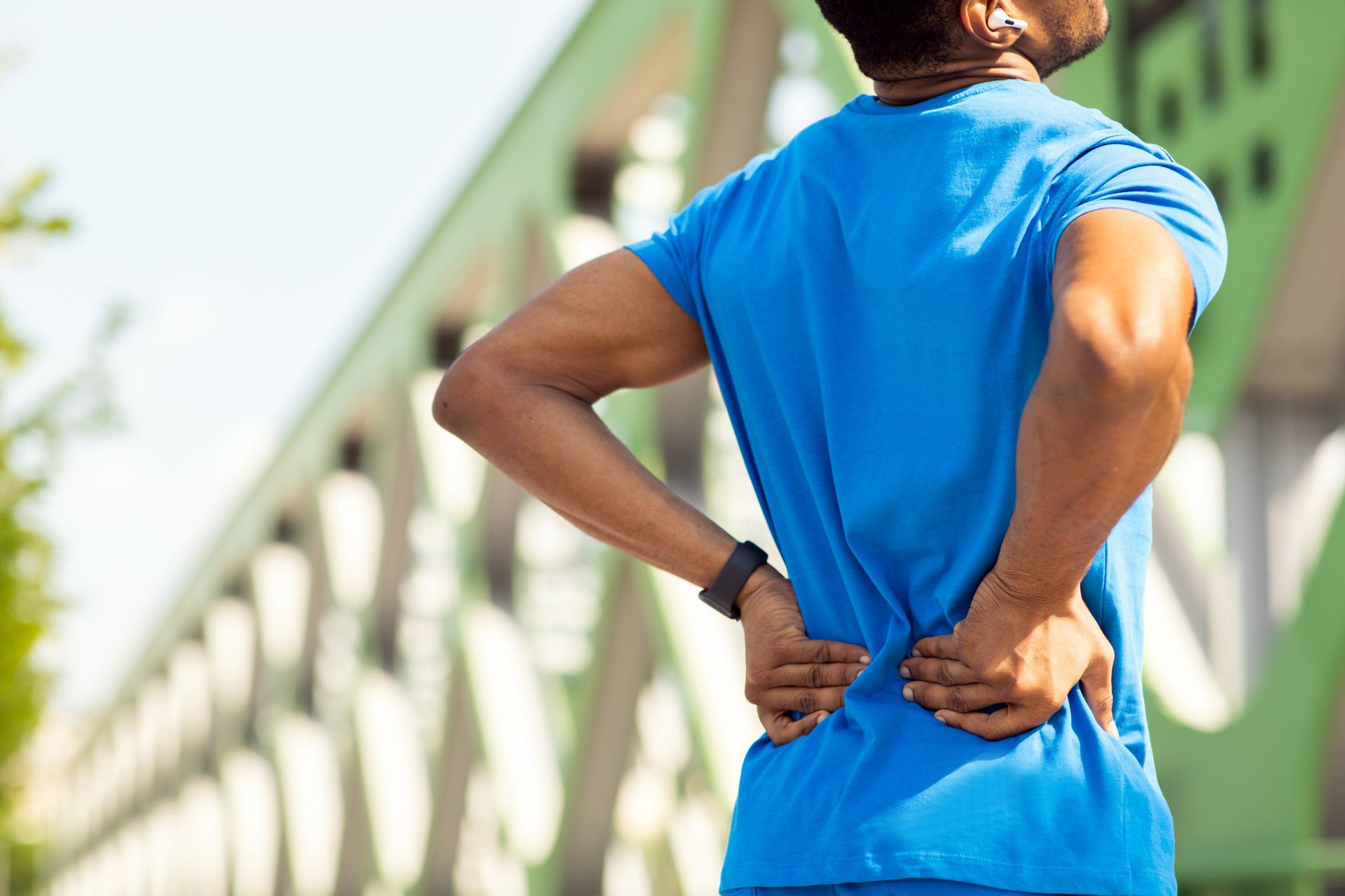 A person in a blue shirt standing outdoors, holding their lower back with both hands to indicate pain.