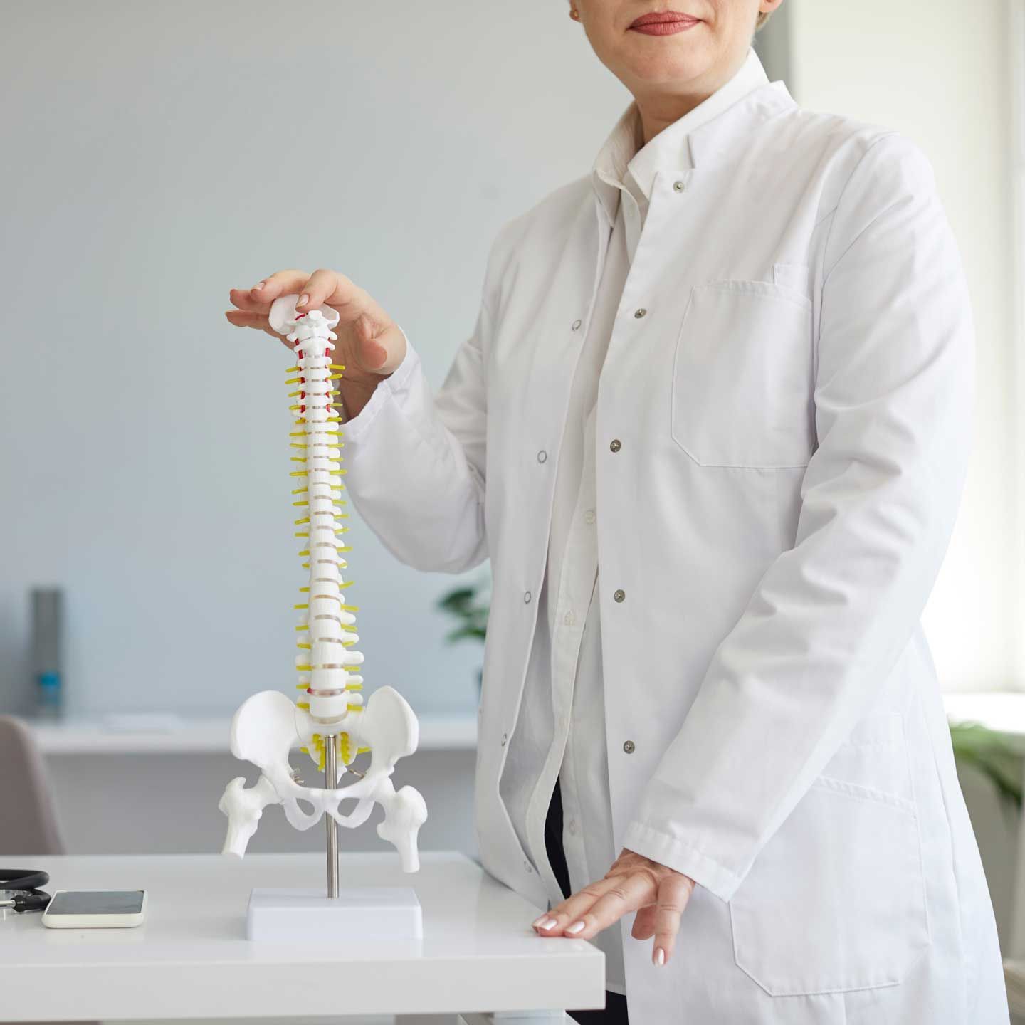 A medical professional in a white coat stands in a clinic, holding a model of a human spine and pelvis.