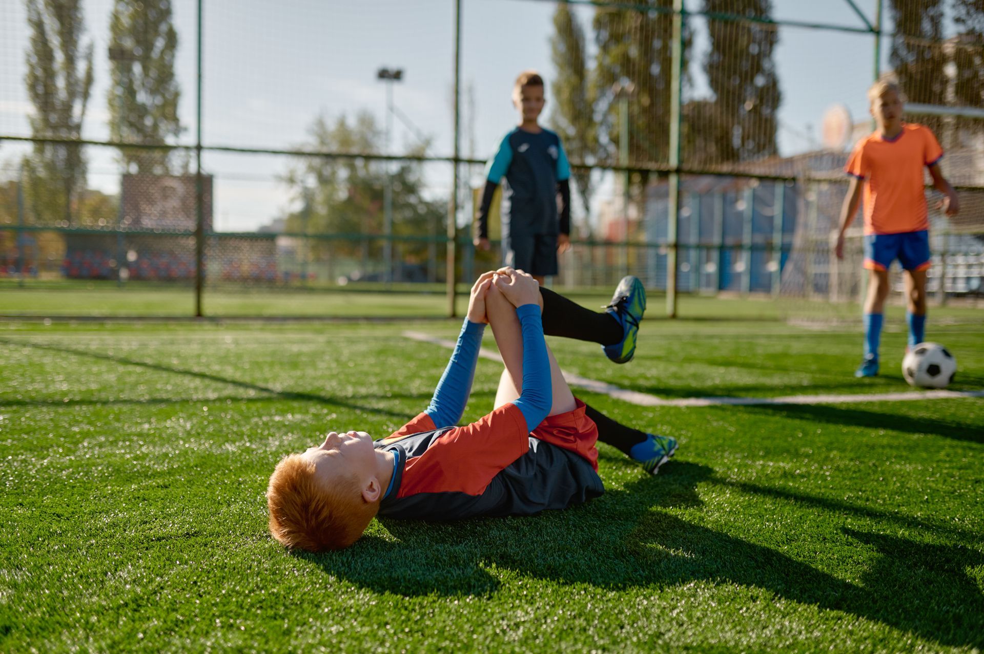 A red-haired child lies on a soccer field clutching their knee in pain as two other children look on nearby.