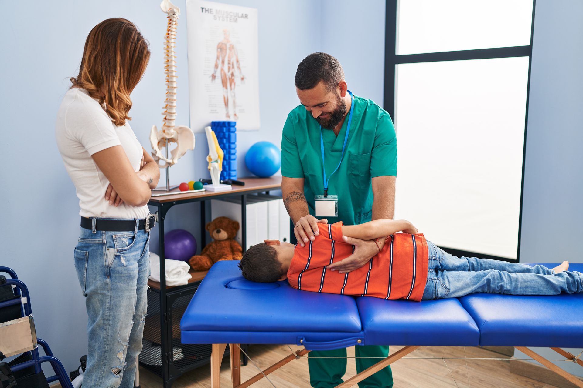 Family having physiotherapy session massaging child back at rehab clinic. A healthcare professional performs physical therapy on a child lying on a treatment table while a parent watches.