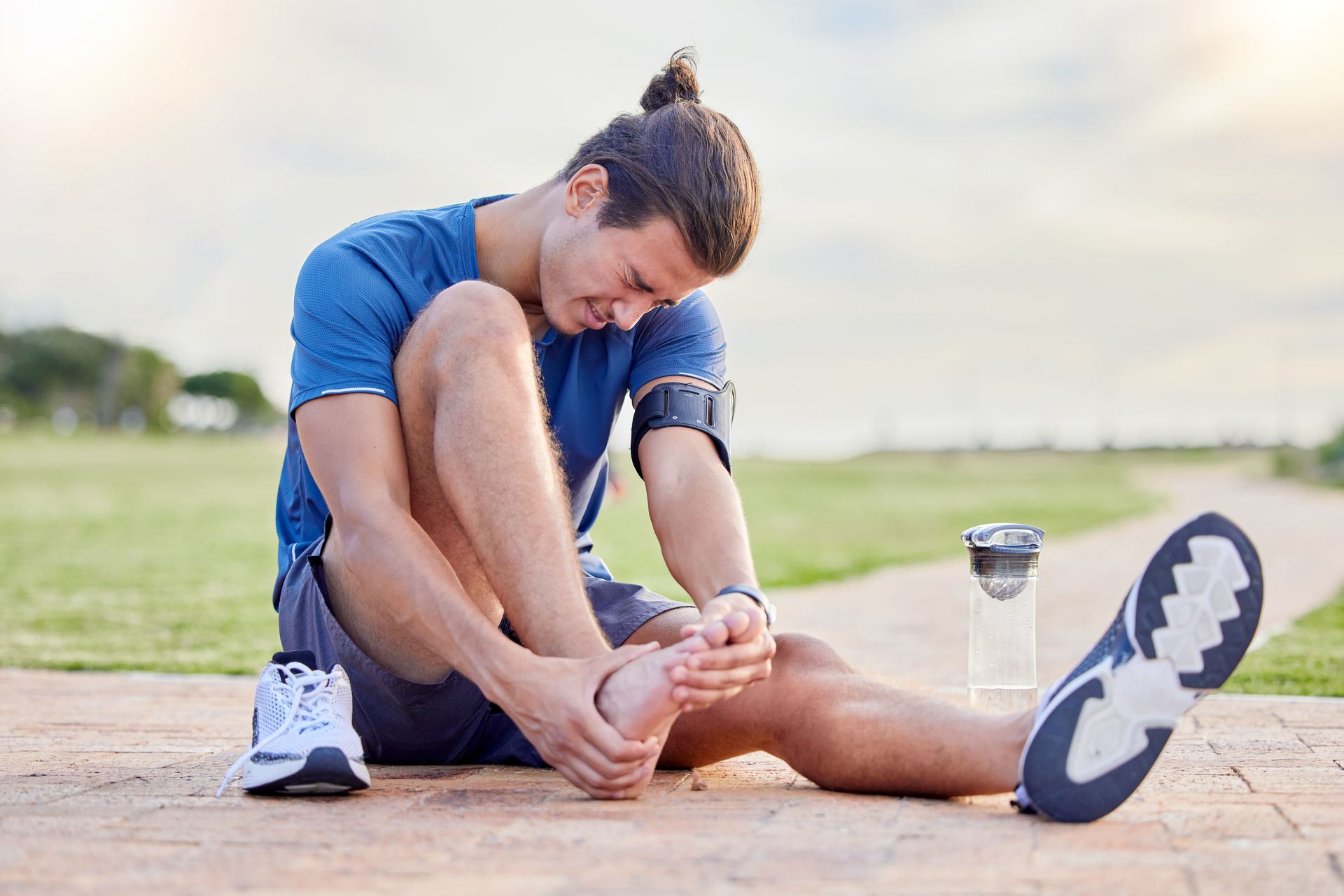 A runner sits on an outdoor path, holding their foot and wincing in pain, with one shoe removed.