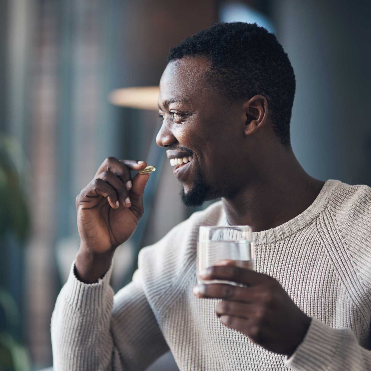 A person smiling while preparing to take a supplement pill with a glass of water in a softly lit, modern setting.
