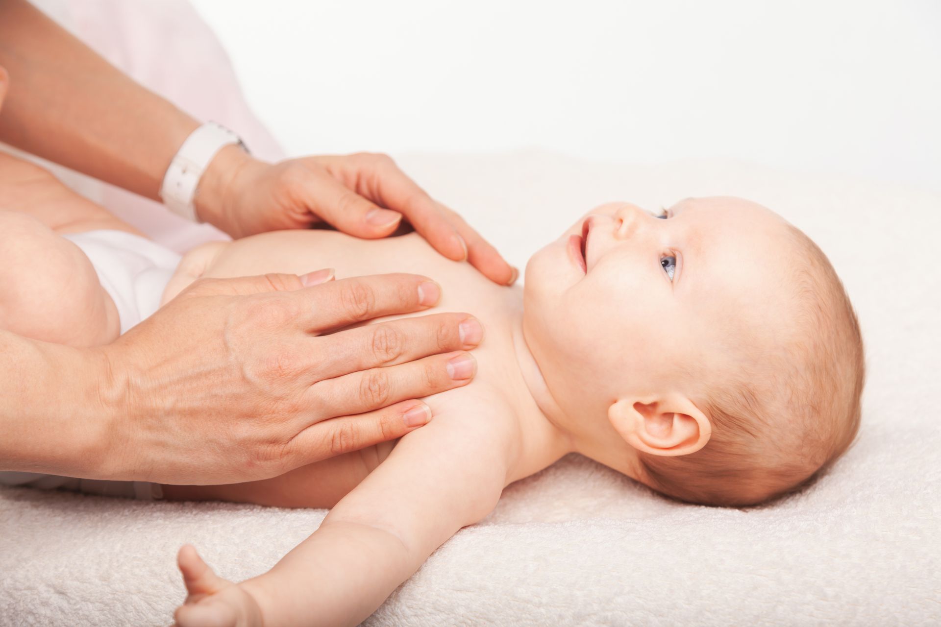 An adult's hands gently massage the chest of a baby lying on a soft, white surface.