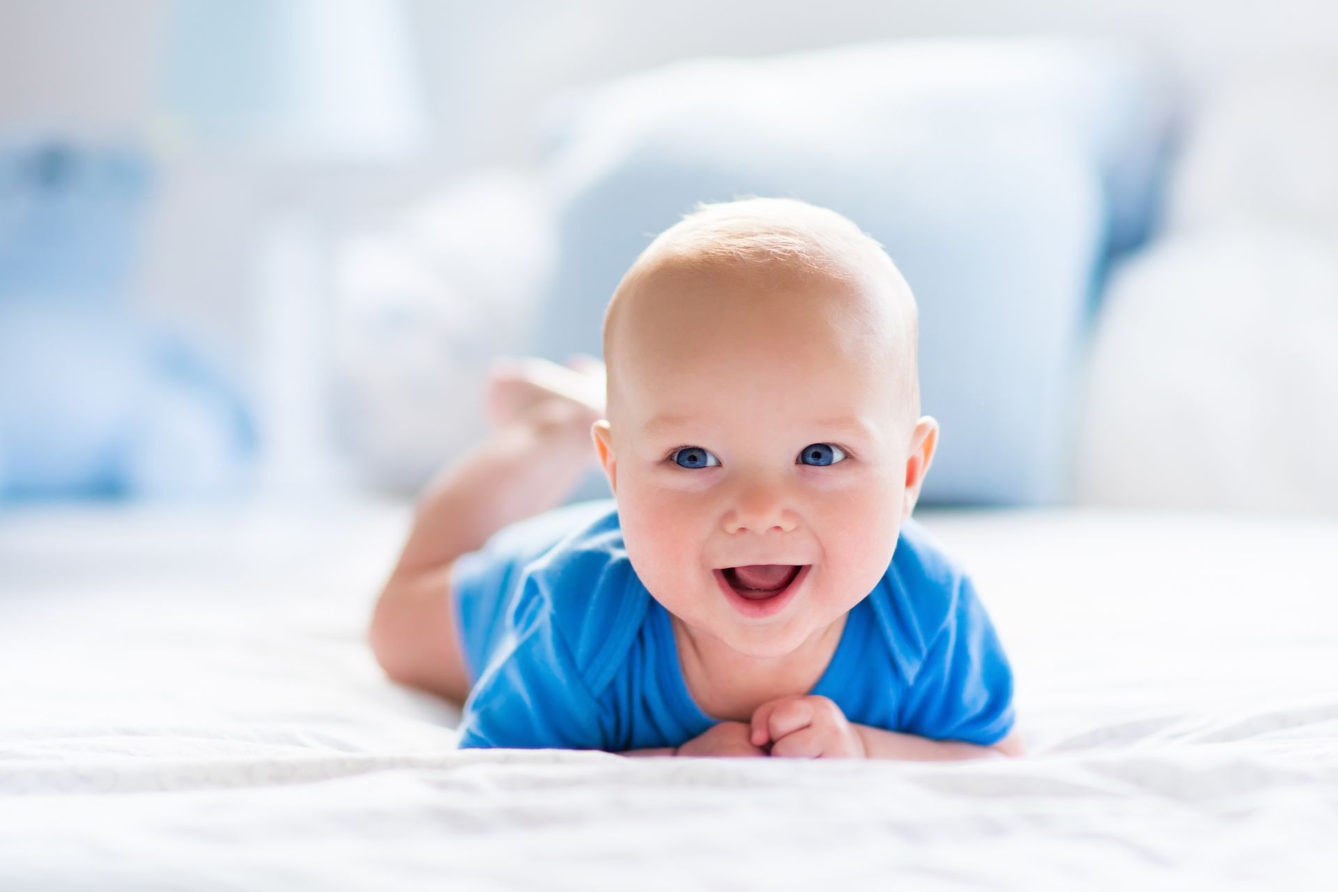 A smiling baby wearing a blue shirt lies on their stomach on a white bed.