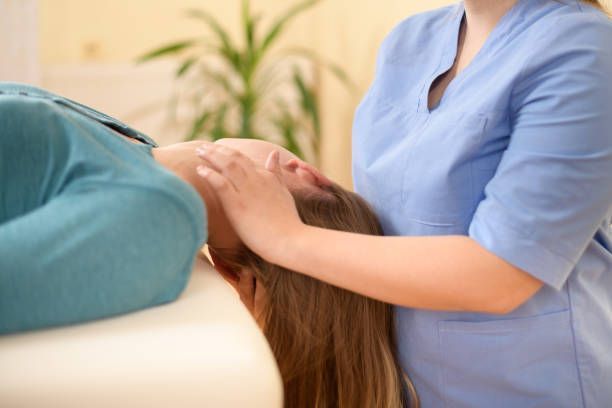 A person in blue scrubs performs a gentle massage on the temples and forehead of a client lying on a massage table.