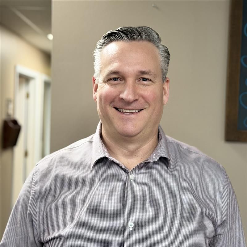 A smiling professional wearing a grey button-down shirt stands in a bright, neutral-toned office space.