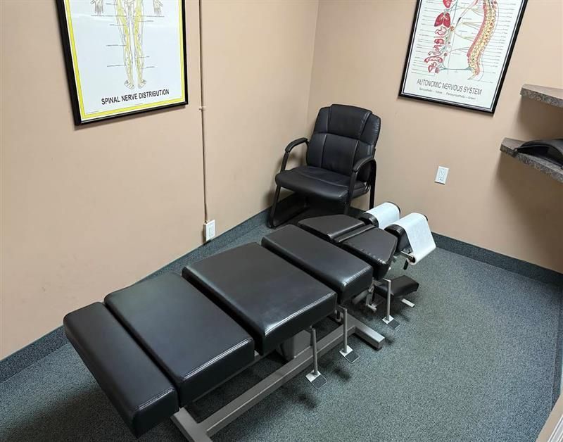 Person pointing to a spinal model, highlighting the lumbar region. A brown chiropractic treatment table stands in a corner office with a black chair and medical posters on beige walls.