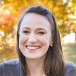 Woman with brown hair smiles in front of fall foliage.