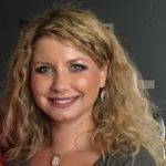 Blond woman with curly hair smiles, wearing a gray shirt and necklace.