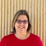 Woman with glasses smiles in front of a wooden wall, wearing a red shirt.