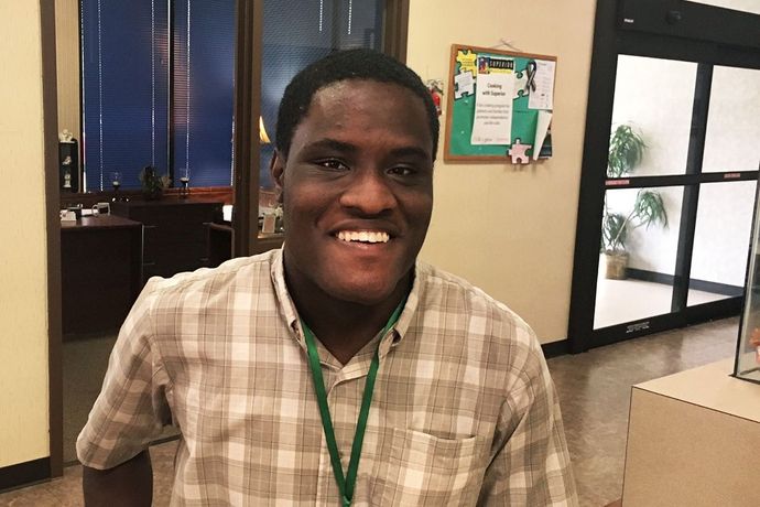 Smiling Black man wearing a plaid shirt and lanyard stands indoors near a bulletin board and glass doors.