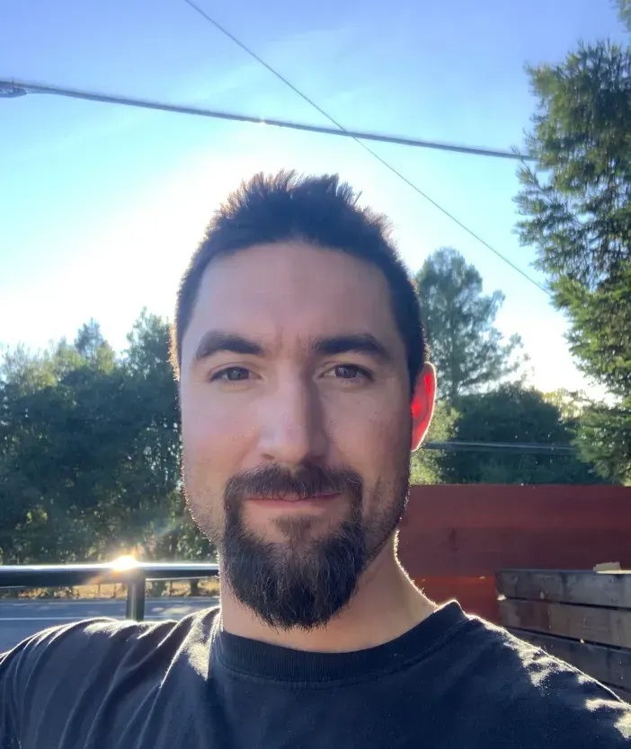 Man standing outdoors in sunlight with trees and a blue sky in the background