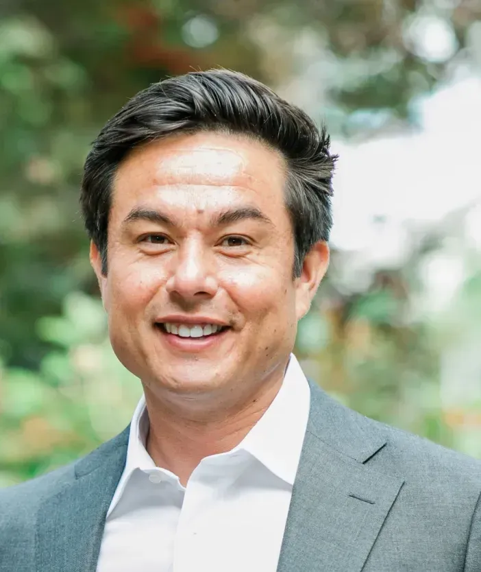 Smiling man in a gray blazer and white shirt outdoors with blurred greenery behind him