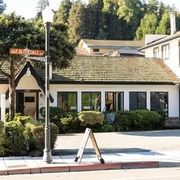 Small white cottage-style storefront with a shingled roof, streetlamp, sign, and trees in the background.