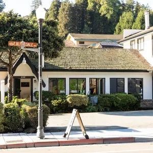 Small white cottage-style storefront with a shingled roof, streetlamp, sign, and trees in the background.