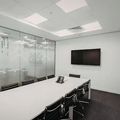Modern conference room with a long white table, black chairs, glass walls, and a wall-mounted TV.