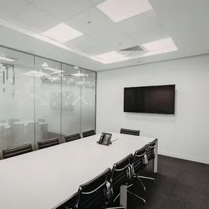 Modern conference room with a long white table, black chairs, glass walls, and a wall-mounted TV.