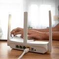 Hands typing beside a white Wi‑Fi router with antennas on a wooden desk