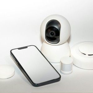 White security camera beside a smartphone and smart home devices on a white surface