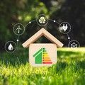 Small wooden house with energy efficiency label, surrounded by green grass and renewable energy icons on a blurred background