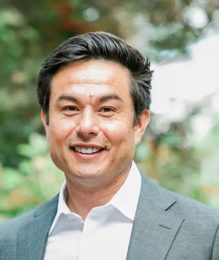 Smiling man in a gray blazer and white shirt outdoors with blurred greenery behind him