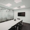 Modern conference room with a long white table, black chairs, glass walls, and a wall-mounted TV.