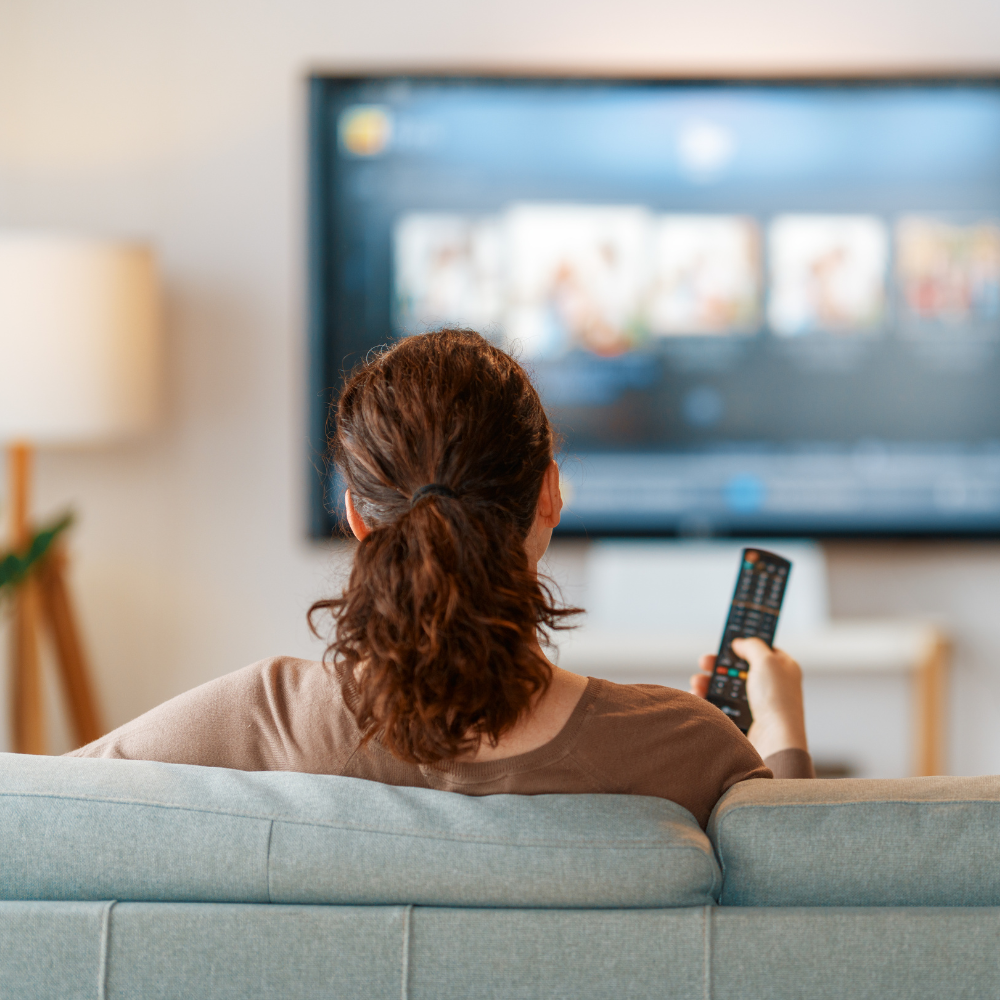 Person on a couch holding a remote, watching a TV in a softly lit living room.