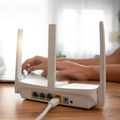 Hands typing beside a white Wi‑Fi router with antennas on a wooden desk