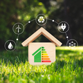 Small wooden house with energy efficiency label, surrounded by green grass and renewable energy icons on a blurred background