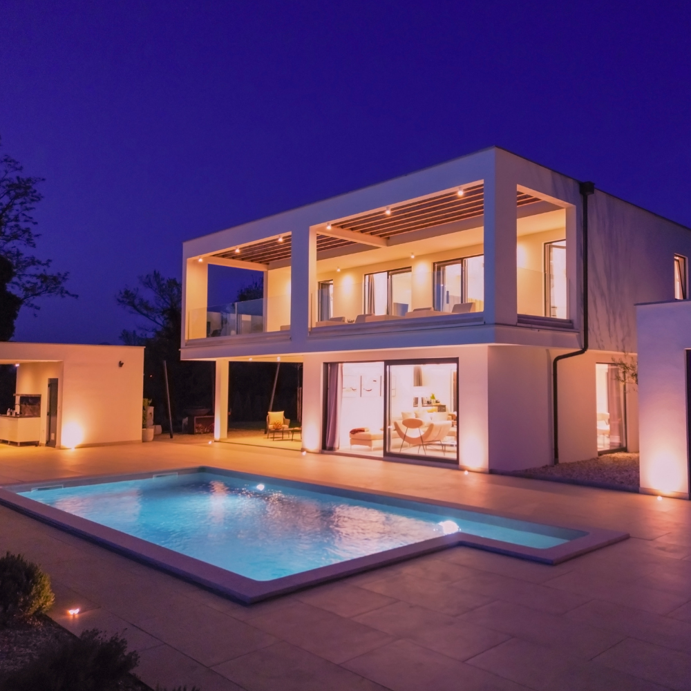 Modern two-story house at dusk with lit terrace and a glowing blue pool in the foreground