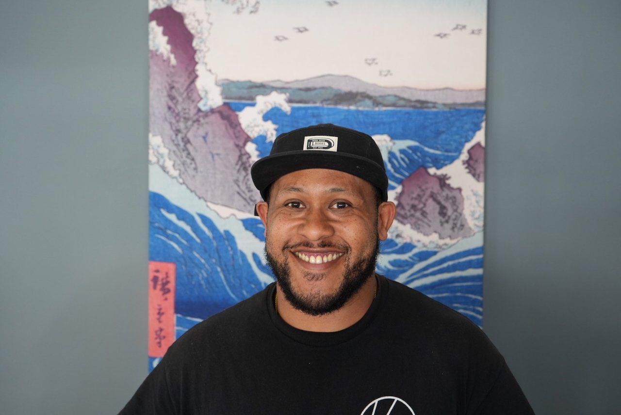 Smiling person in black cap and shirt standing before a colorful ocean-wave painting