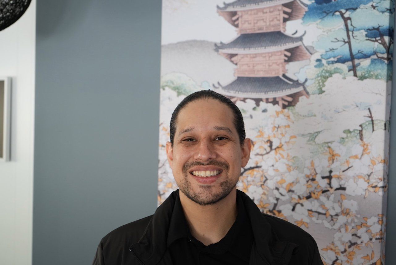 Smiling person in a black shirt standing indoors before a wall mural with a pagoda and cherry blossoms