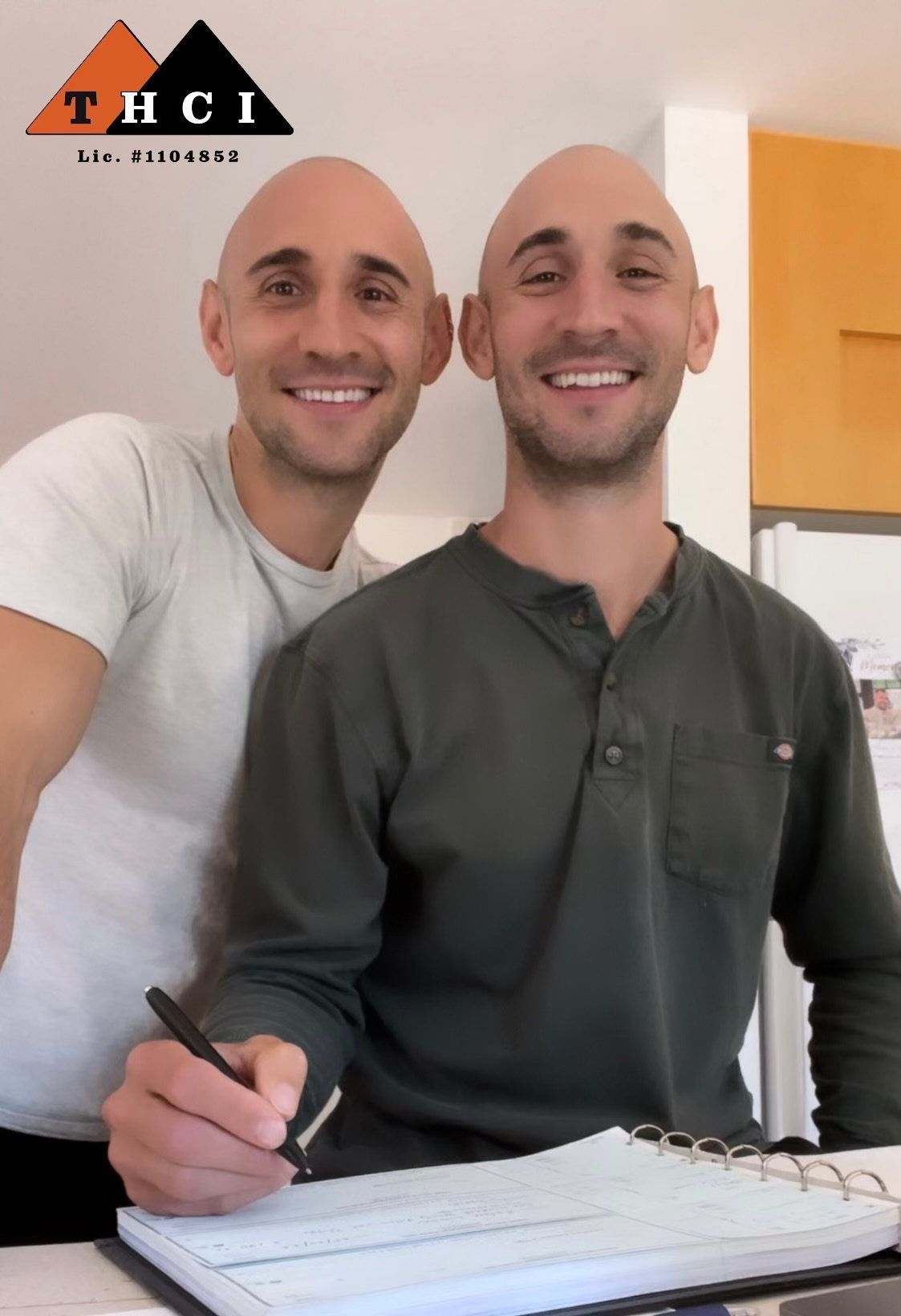 Two smiling people sitting at a table, one writing in a notebook, indoors beside an orange wall.