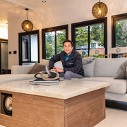 Man seated on a gray sofa in a modern living room with large windows and a wooden coffee table