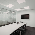 Modern conference room with a long white table, black chairs, glass walls, and a wall-mounted TV.