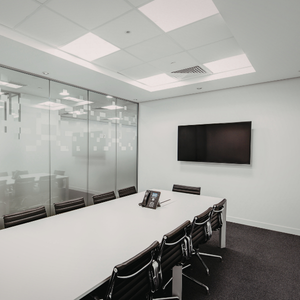 Modern conference room with a long white table, black chairs, glass walls, and a wall-mounted TV.