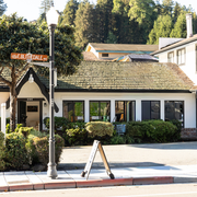 Small white cottage-style storefront with a shingled roof, streetlamp, sign, and trees in the background.
