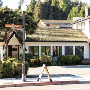 Small white cottage-style storefront with a shingled roof, streetlamp, sign, and trees in the background.