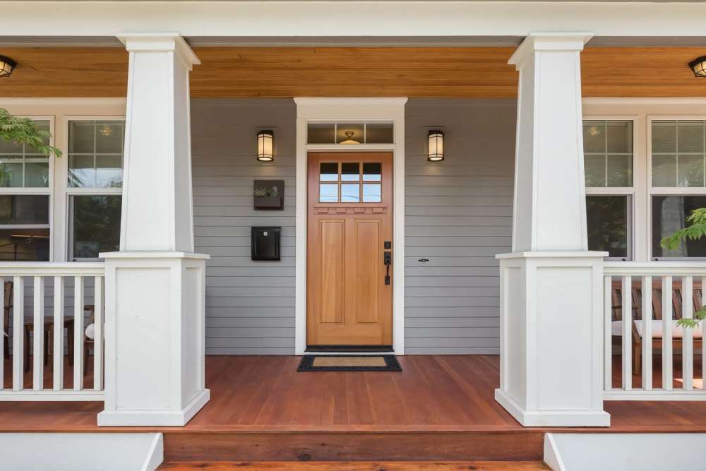 Front porch of a house with light gray horizontal siding, a natural wood door, white columns, and wood decking.