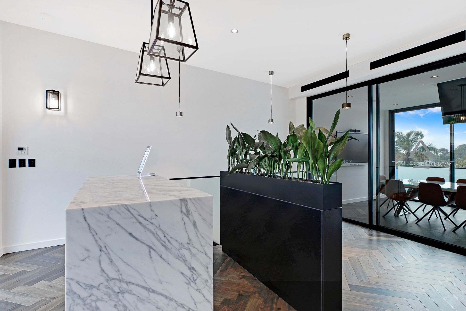 A marble island and black planter with green plants stand in a modern, white-walled room with herringbone floors. — Trim Joinery In Fountaindale, NSW