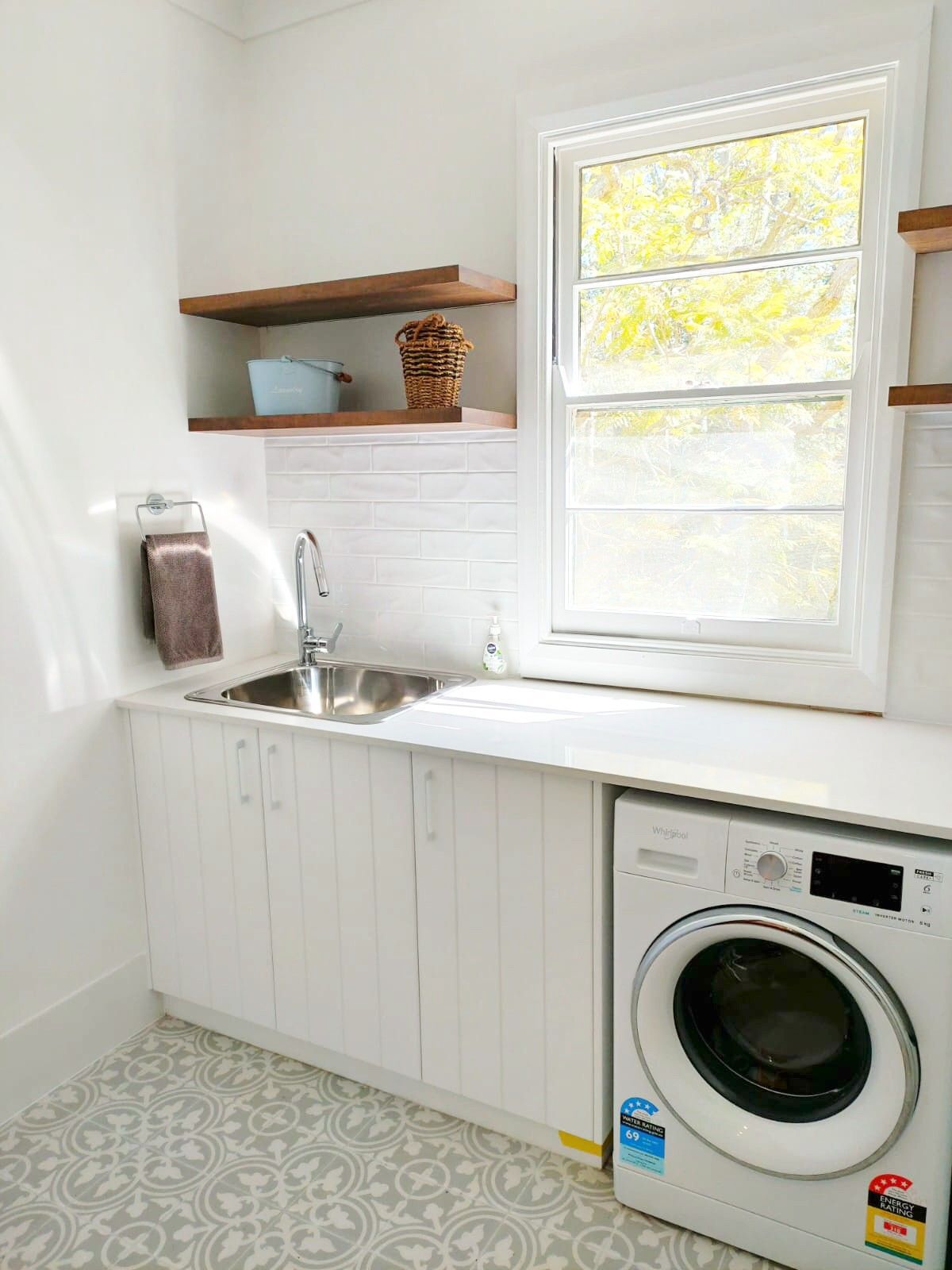 A bright laundry room with white cabinets, a sink, wooden floating shelves, a front-load washer, and patterned floor tiles. — Trim Joinery In Fountaindale, NSW