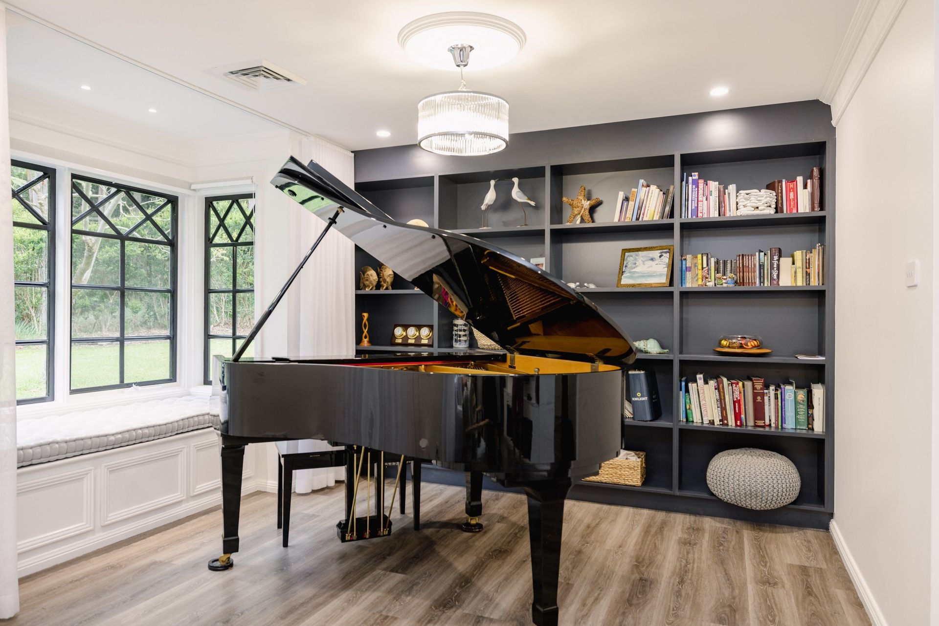 A grand piano sits in a room with light wood floors, a bay window, and a dark built-in bookcase filled with books. — Trim Joinery In Fountaindale, NSW
