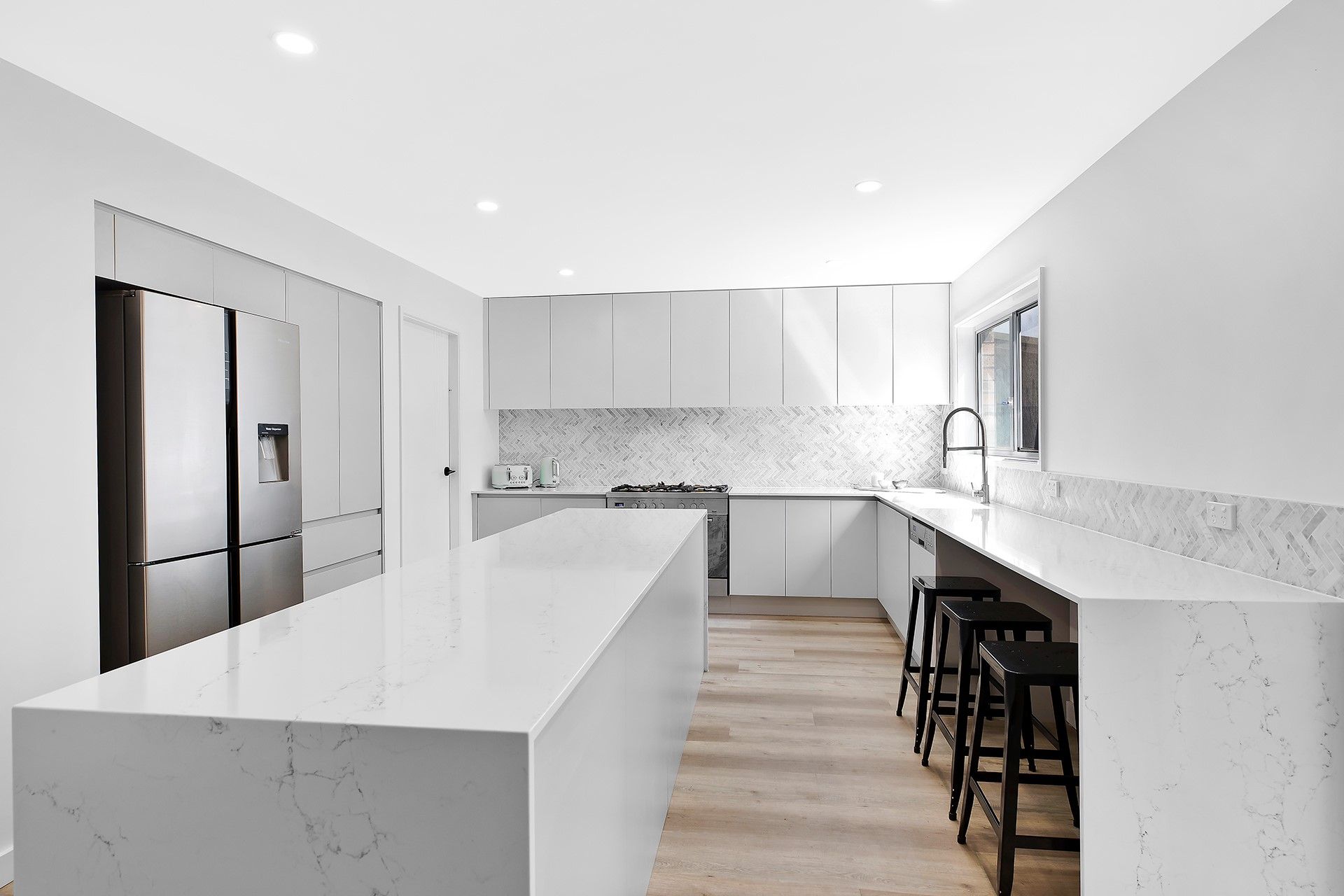 A modern, white kitchen with a large marble-topped island, stainless steel refrigerator, and bar stools at a side counter. — Trim Joinery In Fountaindale, NSW